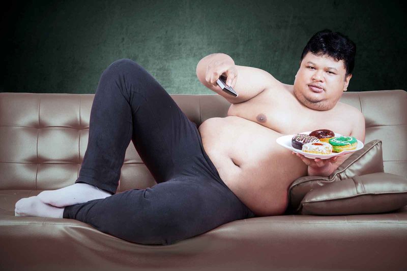 fat man on couch holding TV remote and donuts